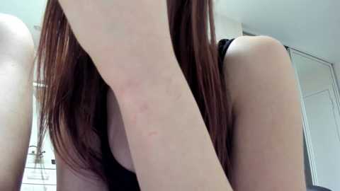 Video of a young woman with long, straight brown hair, wearing a black sleeveless top, standing indoors with pale skin and noticeable red marks on her right arm.