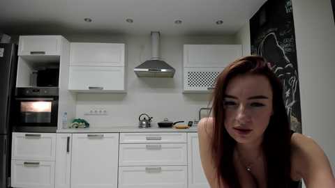 Video of a modern, white kitchen with a young woman with long red hair and light skin, wearing a black top, standing in the foreground.