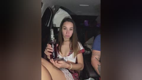 Video of a young woman with long brown hair, wearing a plaid skirt and white top, holding a bottle of vodka, sitting in the backseat of a car.