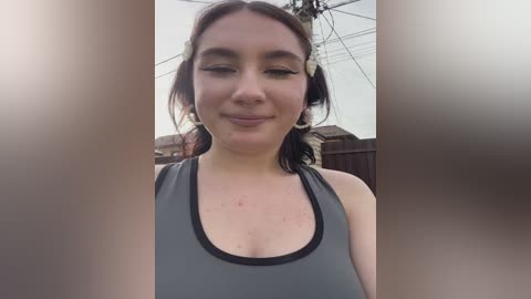 A video of a fair-skinned woman with shoulder-length brown hair, wearing a gray sports bra, smiling slightly. She stands outdoors in a suburban setting with a wooden fence and power lines in the background.