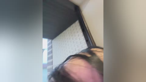 A video of a person with short, dark hair, partially cropped, standing in a room with a white brick wall and a dark, vertical window frame. The lighting is soft, creating a subtle shadow.