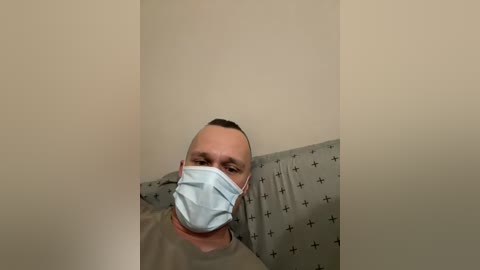 Video of a bald man wearing a light blue surgical mask, lying in a hospital bed with a beige pillow and a gray, patterned blanket. The background is plain beige walls.