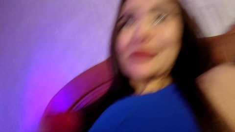 A blurred video of a woman with fair skin and long brown hair, wearing a blue top, lying on a red cushion, surrounded by a purple and pink glow.