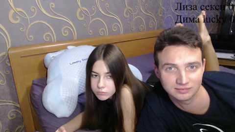 Video of a young couple in a bedroom. The woman, with long brown hair, lies on a bed, looking serious. The man, with short brown hair, sits beside her, smiling. Background shows a wooden headboard, a white pillow, and patterned wallpaper.