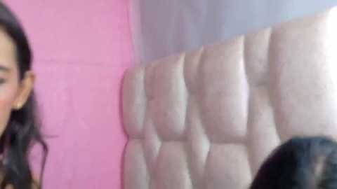 A blurred video of a woman's face with long black hair partially visible. She is standing in a room with a pink wall and a tufted white headboard in the background.