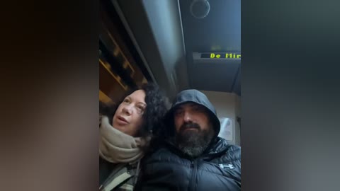 Video of two people, an Asian woman with curly hair in a beige scarf and a bearded man in a dark hooded jacket, sitting close together on a train, with a \"Be Quiet\" sign overhead.