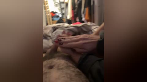 A blurred video of a person lying on a bed, with their head covered by a hand, in a dimly lit, cluttered room with clothes and luggage visible in the background.