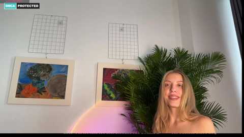 Video of a topless blonde woman with fair skin, smiling, standing beside a green potted plant and colorful abstract paintings on a white wall.