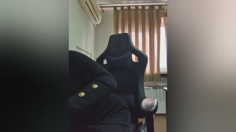 Video of a dark-skinned person sitting in a black gaming chair, arms raised, wearing a black jacket, in a dimly lit room with beige walls, brown curtains, and an air conditioner.