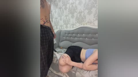 Video: A tattooed woman in plaid pants stands over a bald, tattooed man lying on a bed, wearing a blue bra. Background: Gray bed with floral wallpaper.