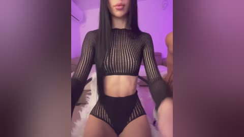 Video of a slender, light-skinned woman with long black hair, wearing a black fishnet crop top and matching high-waisted briefs, sitting on a bed with white sheets and a pillow. Purple lighting casts a soft glow in the background.