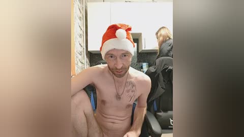 Video of a topless white man with a red Santa hat and a tattoo on his chest, sitting in a modern kitchen with a woman in the background.