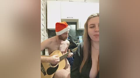 Video of a shirtless man with a red Santa hat playing an acoustic guitar in a modern kitchen, partially obscured by a blurred figure in the foreground.