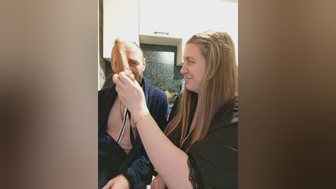 A video captures a woman with long, straight hair being styled by a woman in a black robe in a modern kitchen. The background features white cabinetry and dark countertops.