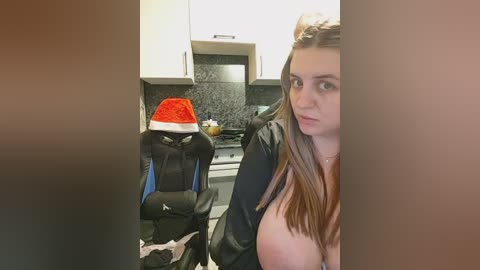 Video of a young woman with light skin and long brown hair, wearing a black jacket, sitting in a modern kitchen with a red Santa hat on a gaming chair, and a stove in the background.