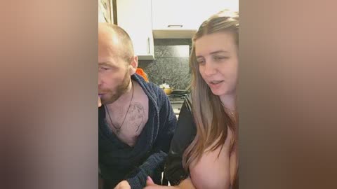 A video captures a couple in a kitchen. The man, bald, with a beard, is shirtless, revealing a tattoo. The woman, with long brown hair, is topless, exposing her breasts. Both appear to be engaged in a passionate moment.