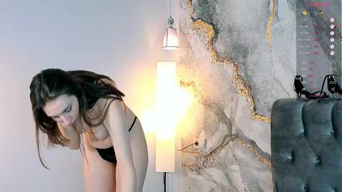 Video of a slender Caucasian woman with long brown hair, wearing black lingerie, bending forward in a minimalist room with a textured grey wall, a lit floor lamp, and a grey upholstered chair.