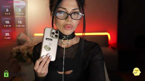 Video of a woman with glasses and a black collar, holding a phone, in a dimly-lit room with red lighting, wearing a black sheer top.