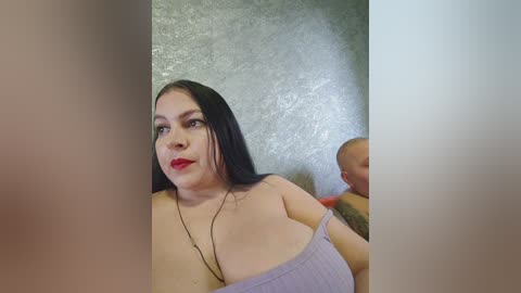 Video of a fair-skinned, dark-haired woman with red lipstick, wearing a lavender off-shoulder top, holding a baby with a bald head, against a textured gray wall.