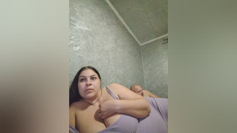 Video of a plus-size woman with long black hair, wearing a lavender ribbed top, lying on her side in a dimly lit room with textured green walls. Her expression is neutral.