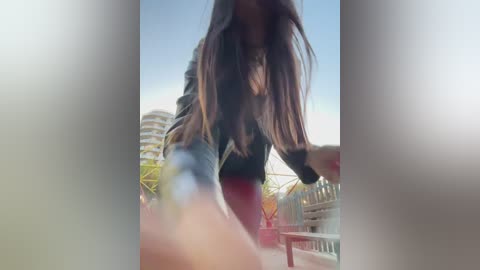 Video of a woman with long brown hair, wearing a black top and red pants, taken from a low angle. Background features a modern building and clear sky, creating a bright, urban setting.