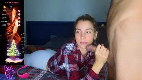 A video shows a young woman with light skin and brown hair, wearing a plaid shirt, performing oral sex on a naked man. The bedroom background includes a bed with a blue headboard.