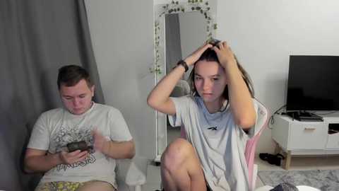 Video of a man in a gray shirt and shorts, and a woman in a white t-shirt, sitting on a pink chair in a living room with a TV, mirror, and green wall decor.