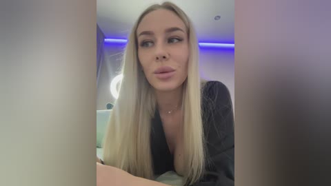 Video of a blonde woman with long hair, wearing a black top, making a pouty face, in a dimly lit room with blue lighting.