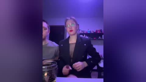 Video of a woman with red lipstick and glasses in a black blazer, sitting next to a man in a grey shirt at a dimly lit bar.