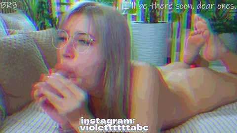 A blurred video of a young woman with fair skin, straight blonde hair, and glasses, lying nude on a couch, eating a popsicle, surrounded by colorful potted plants.