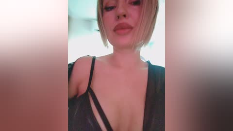 A video of a fair-skinned woman with short, blonde hair, wearing a black lace dress with a plunging neckline, taken indoors with a soft focus.