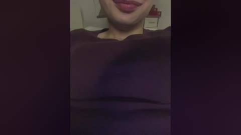 Video of a person with light skin, wearing a dark brown shirt, with a hint of their lips visible. Background shows a blurry nightstand and lamp.