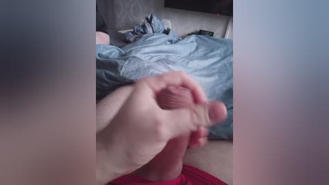 A close-up video of a hand holding a flaccid penis over a bed covered with a blue and white quilt. The background shows a messy room with clothes and a dark TV stand.