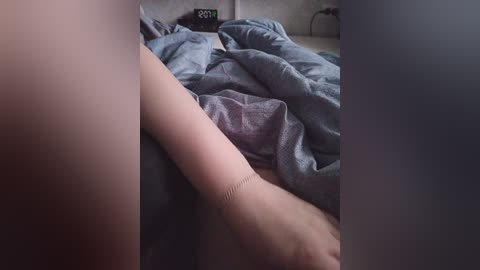 A video showing a person's arm and hand, covered in a grey blanket, lying on a bed with a clock displaying 8:17 on a nightstand in the background.