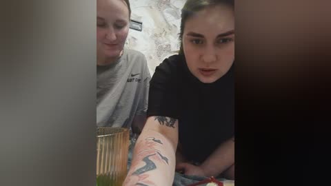 Video: A woman with a black tattooed arm and short hair, wearing a black t-shirt, looks at a woman with a grey Nike shirt, sitting at a table.