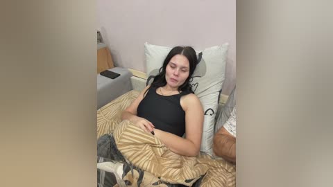 A video shows a young woman with long black hair, wearing a black tank top, lying on a bed with beige patterned bedding, surrounded by pillows and a beige wall.