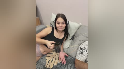 A young woman with long dark hair and fair skin, wearing a black top and pink panties, sits on a bed, holding a coffee cup, in a dimly lit room.