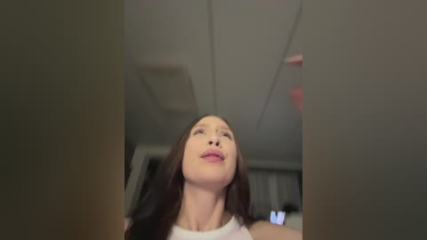 A video of a woman with long, dark hair and light skin, wearing a white top, captured from a low angle, looking upwards with a serene expression. The background is dimly lit, showing a ceiling and blurred furniture.