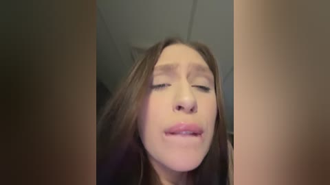 A close-up video of a young woman with light skin and straight brown hair, eyes closed, mouth slightly open, and a soft expression. The background is blurred, suggesting an indoor setting with a ceiling.