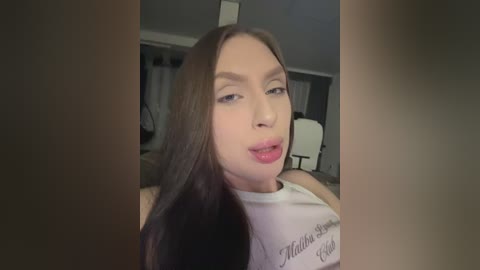 Video of a young woman with long brown hair, light skin, and pink lipstick, wearing a white tank top with text, smiling slightly. Background shows a dimly lit room with a chair and window.