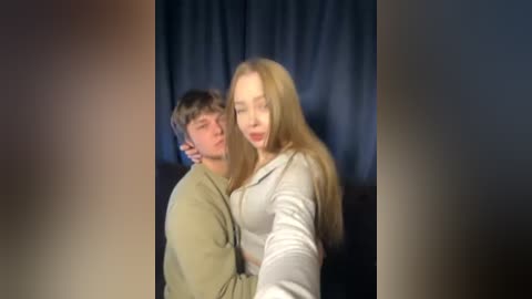 Video of a young couple, a fair-skinned man with short brown hair, and a fair-skinned woman with long blonde hair, both smiling, standing close, in a dimly lit room with blue curtains.