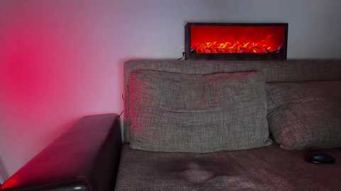 A video of a beige fabric sofa with a fire effect LED display mounted above it, emitting red light. The background wall is plain white, and a small black remote control lies on the sofa.
