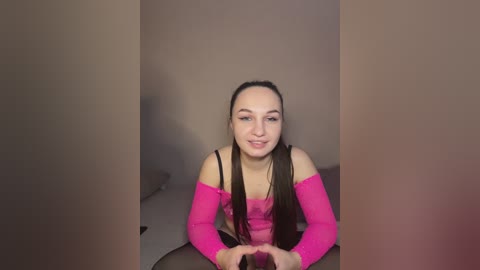 Video of a smiling young woman with fair skin and straight black hair, wearing a pink off-shoulder top and black leggings, sitting on a gray couch against a beige wall.