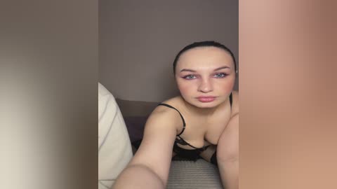 Video of a light-skinned woman with dark hair, wearing black lingerie, lying on a bed with beige sheets.