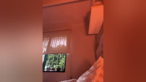 A video of a dimly-lit, small room with beige walls, a window with a partially open white shade, and a bed with white sheets. The room has a warm, orange glow, suggesting artificial light.