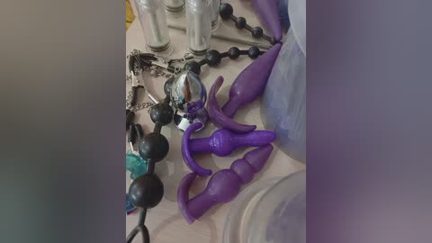 Video of a messy table with a black and purple assortment of BDSM sex toys, including balls, handcuffs, and dildos, with a metallic chain and a clear plastic bag in the background.