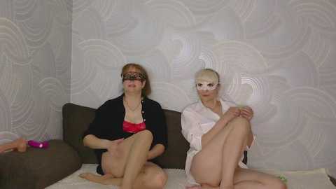 Video of two elderly women with fair skin, wearing black blindfolds, sitting on a beige sofa in a room with white, wavy-patterned wallpaper. One wears a black robe, the other a white one.