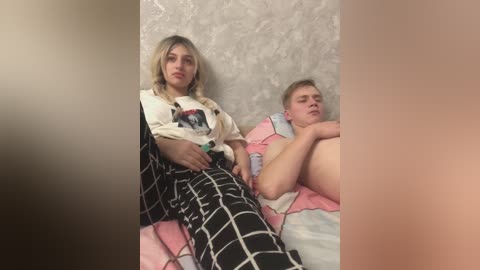 A video shows a blonde woman in a plaid skirt, sitting beside a shirtless, fair-skinned man, both on a bed with a patterned blanket. The background is a textured, light gray wall.