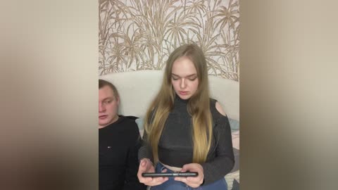 Video of two young women seated on a couch, one with long blonde hair in a grey top, the other with short brown hair in a black hoodie, both looking at a tablet. Background shows beige wallpaper with palm tree pattern.
