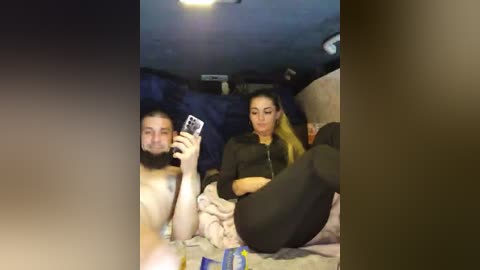 Video of a man with a beard and a woman with long hair, both topless, sitting on a bed. The background features dim lighting and a few scattered items.